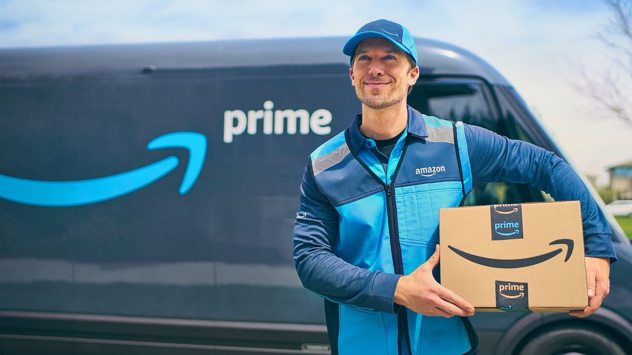 An Amazon Prime delivery driver in a branded cap and vest holds a Prime-taped cardboard box in front of a Prime delivery van.