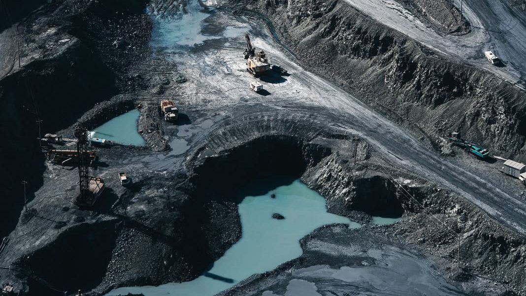 Aerial view of a terraced cobalt open-pit mine in the Democratic Republic of Congo, its black rock benches cut by pale haul roads, with an excavator, haul trucks and a drill rig working beside pools of turquoise water.