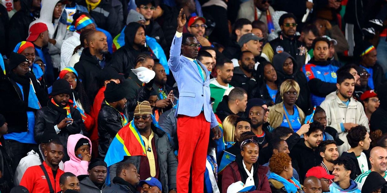 A supporter in a light blue blazer stands above a stadium crowd with his arm raised, surrounded by fans wrapped in Democratic Republic of Congo flags.
