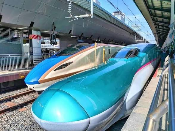 Two Shinkansen bullet trains, one blue and gold and one teal, standing side by side at a Japanese railway platform.