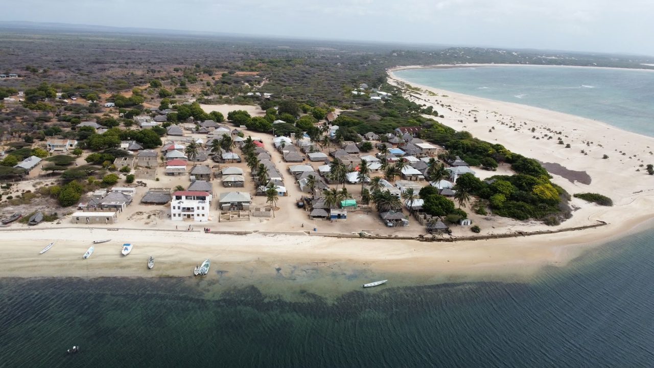 Aerial view of a coastal village in Lamu County on a narrow sand spit, its palm-shaded tin and thatch roofs between a wide empty beach on one side and shallow turquoise water with moored skiffs on the other.