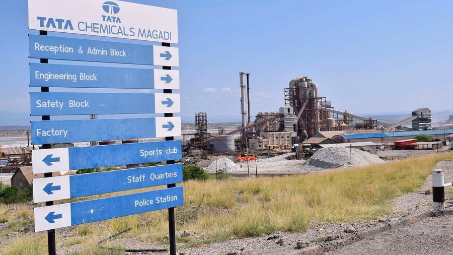 A blue and white signpost reading TATA Chemicals Magadi, directing visitors to the reception, engineering, safety and factory blocks, with the soda ash plant and its chimneys behind it on dry grassland.