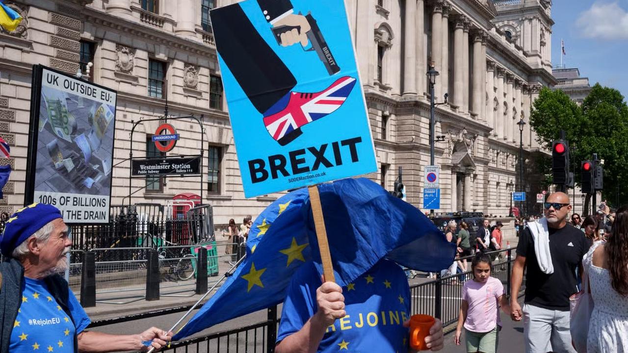 A protester outside Westminster station holds a placard reading BREXIT, beside a banner reading UK outside EU, 100s of billions in the drain. Others nearby wear EU flags and Rejoin EU shirts.