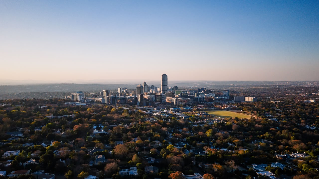 Aerial view of the Sandton skyline at dusk, its cluster of high-rise towers surrounded by low, densely wooded suburbs stretching to the horizon.
