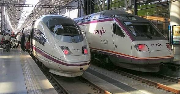 Two Spanish high speed trains, a maroon-striped AVE and a blue-striped Renfe Avant, standing nose to nose on parallel platforms under a glass-roofed station.