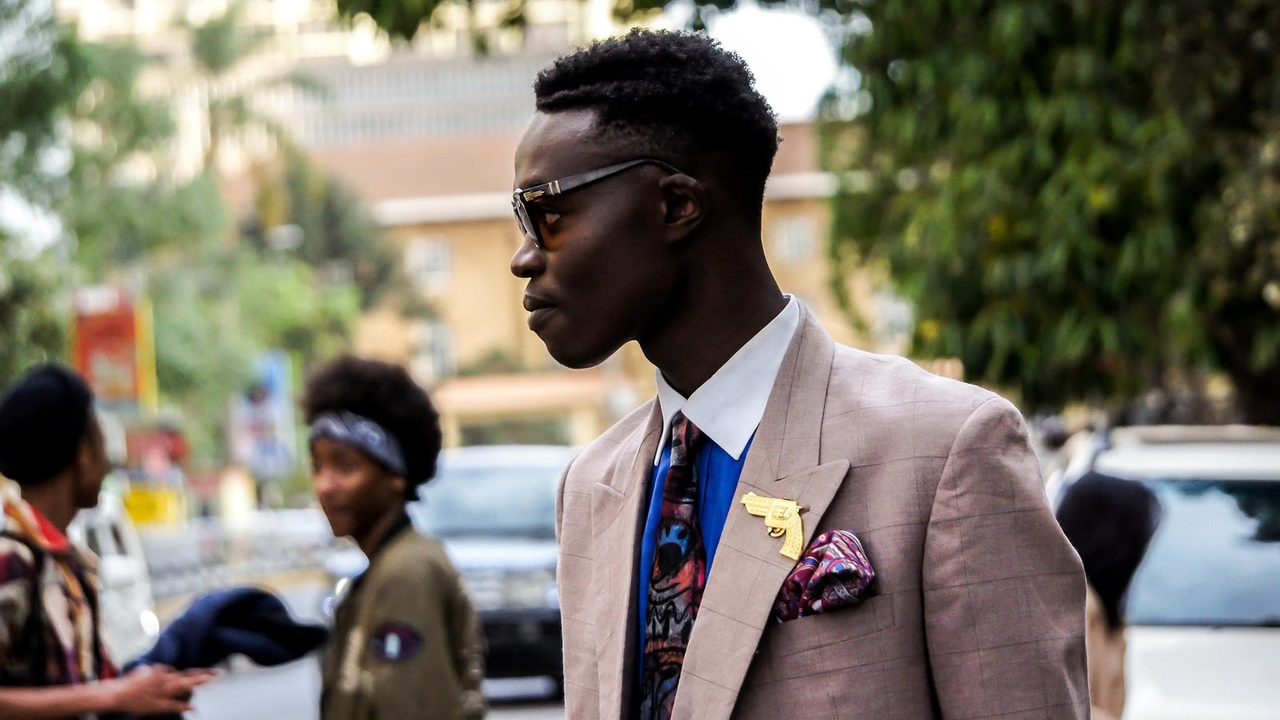 A young man in a checked blazer, open collar and patterned tie stands on a busy street wearing sunglasses and a gold brooch, with other pedestrians out of focus behind him.