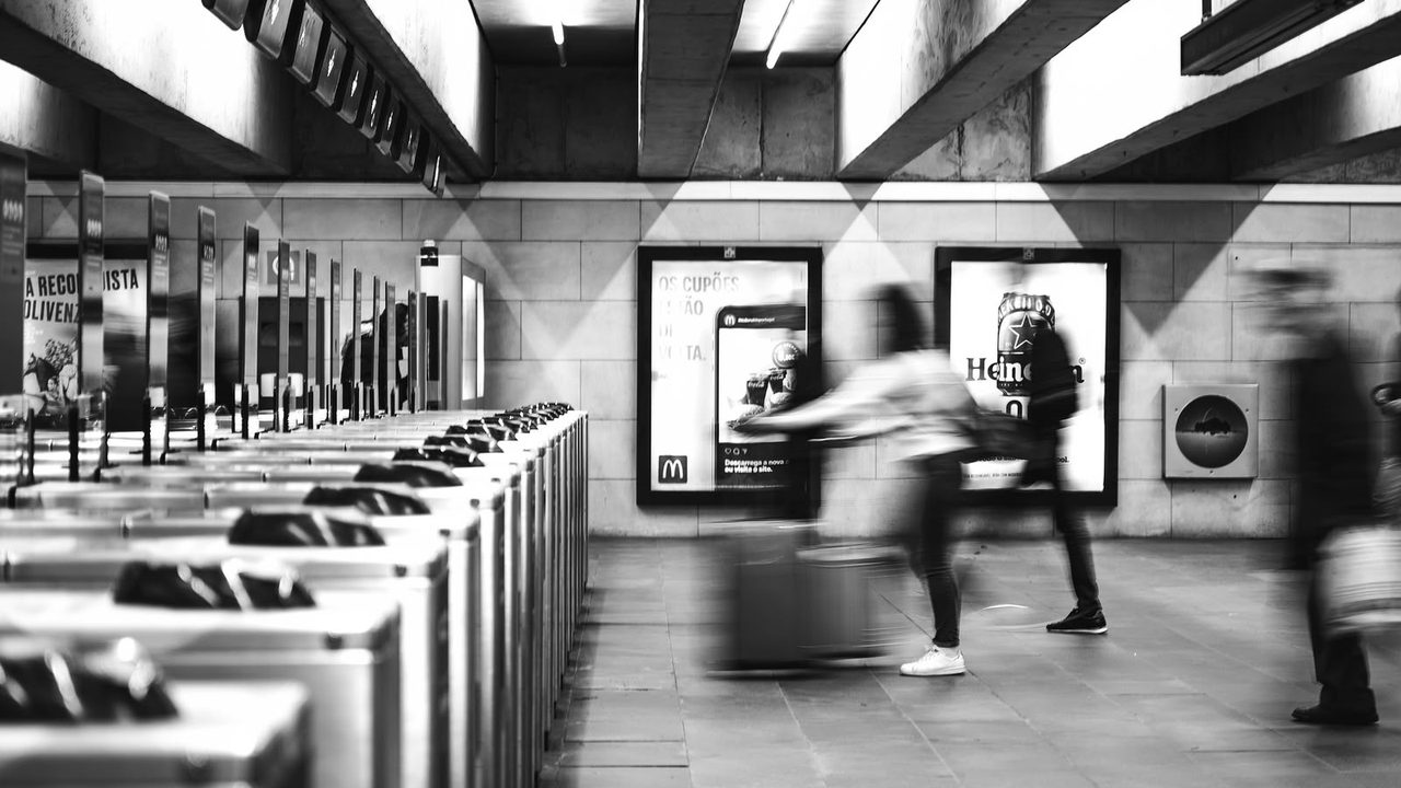 A long row of ticket barriers in a metro station, photographed in black and white, with commuters blurred by motion as they cross the concourse behind.