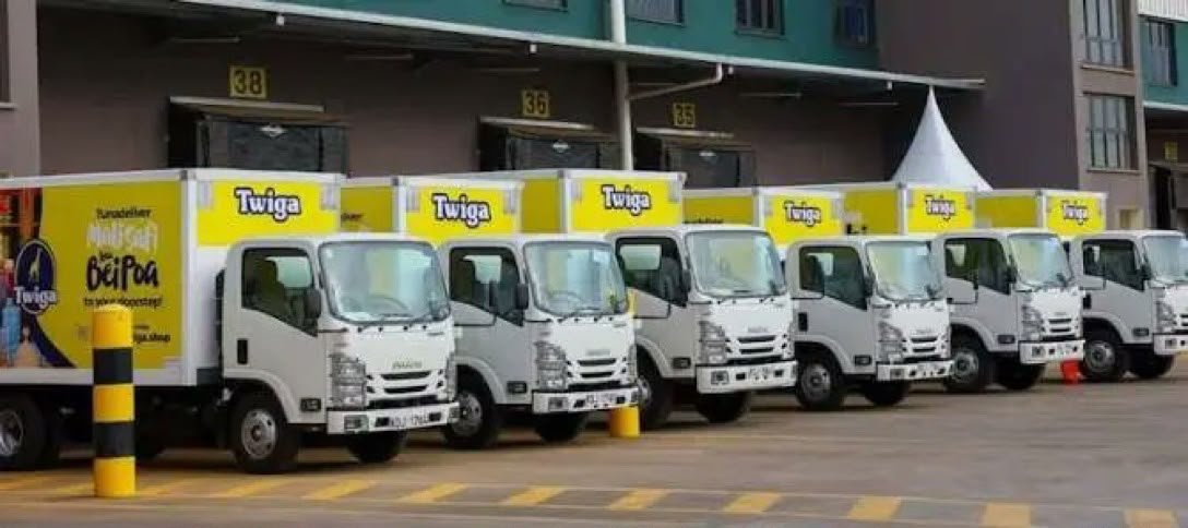 A row of white Twiga-branded delivery trucks parked outside a warehouse, each printed with the Twiga logo and a Swahili delivery slogan.