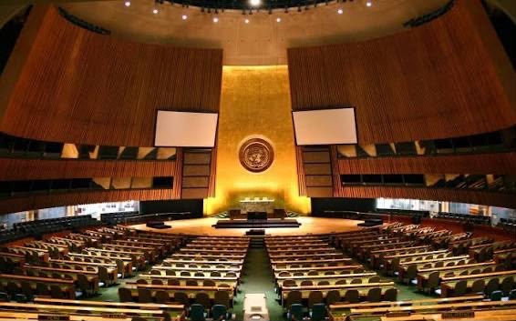 The empty United Nations General Assembly hall, its curved rows of green delegate seats facing the gold-panelled podium beneath the UN emblem.